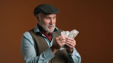 Şapkalı yaşlı bir adam, mavi gömlekli, tüvit yelekli, turuncu arka planda odaklanma ve konsantrasyon gösteren dolarları sayar. Kamera 8K HAM. 