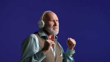 Beyaz sakallı, ritmi hisseden, kulaklıklı müzikten hoşlanan mavi gömlekli, tüvit yelekli, parlak mavi arka planlı son sınıf öğrencisi. Kamera 8K HAM. 