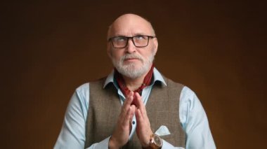 Tüvit yelek ve mavi gömlekli beyaz sakallı yaşlı bir adam, turuncu bir arka plan üzerinde derin bir düşünce jesti olarak ellerini birbirine bağlıyor. Kamera 8K HAM. 