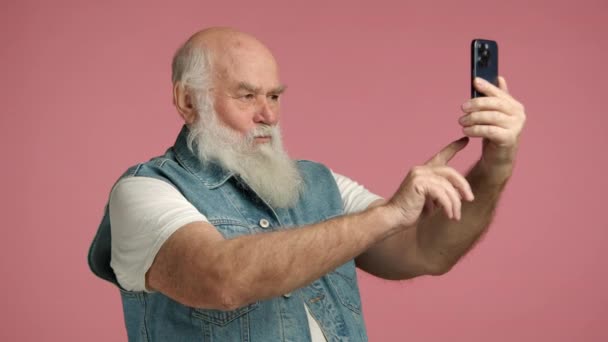 祖父穿着白色T恤 头戴牛仔背心 高兴地拿着智能手机自拍 他笑着为这张照片做鬼脸 祖父秃头 大胡子灰白 — 图库视频影像