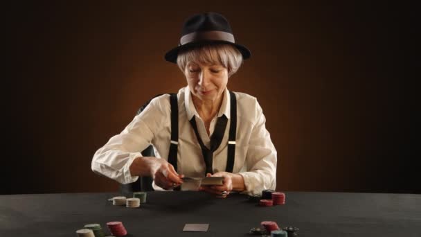 在灯光昏暗的背景下 一位65岁的女士身穿黑社会风格的服装 戴着宽松的领带和黑色的帽子 在扑克游戏中精打细算地打牌 满脸自信 — 图库视频影像