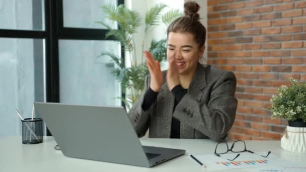 一位穿着西装的年轻职业女性在工作中享受着一个成功的时刻 一边玩乐地跳舞 一边拿着笔记本电脑坐在办公桌前 她的工作空间整洁而现代 略带绿意 — 图库视频影像