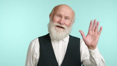 Çekici, yaşlı bir adam dalgayla, ışıl ışıl gözleriyle ve açık ifadesiyle selam veriyor. Çelik yelek ve tişört giymiş, sakin mavi bir arka plan üzerine kurulmuş. Kamera 8K HAM. 