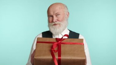 Yaşlı bir adam kırmızı kurdeleli büyük bir hediye kutusu tutuyor, kurnazca gülümsüyor ve deniz mavisi bir zeminde sıcak bir tatil ruhu yayıyor. Kıyafeti zamansız bir zarafeti yansıtıyor. Kamera 8K HAM. 