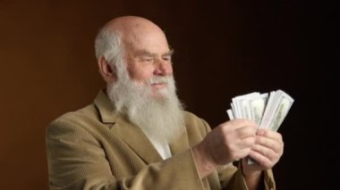 Yaşlı bir adam, bej bir takım elbiseyle sıcacık kahverengi bir arka plana sarılmış bir avuç dolusu nakit parayı sayarken sevinçle ışıldıyor. Onun yüzü emeklilikteki mali güvencenin memnuniyetini yansıtıyor. 8K HAM.