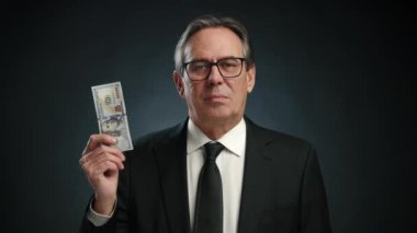 Siyah takım elbiseli bir adam, yüz dolarlık banknotları yırtarken hiçbir duygu göstermez. Belli bağlamlardaki paranın önemsiz değerini simgeler. Kamera 8K HAM. 