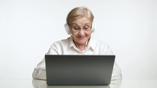 70歳のポジティブな高齢女性は ホワイトバックグラウンド スタジオの上に白いテーブルに座って シルバーラップトップで音楽を聴いています 高品質の4K映像 — ストック動画