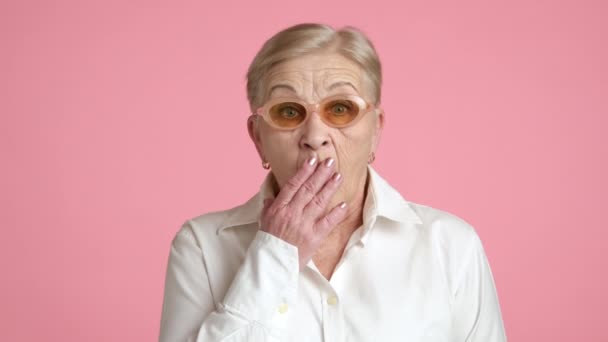 80岁的老妇人戴着时髦的眼镜 白衬衫 用手合上嘴 被吓坏了 站在工作室的粉红背景上 坏消息 高质量的4K镜头 — 图库视频影像