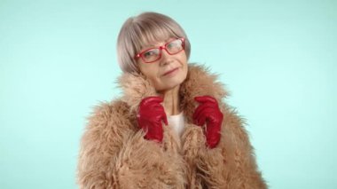 60 'lı yaşlardaki dengeli bir kadın, kışın şıklığını, bronz, sahte kürk manto, kırmızı eldivenler ve gözlüklerle pastel mavisi arka planda koordine eder. Kamera 8K HAM.