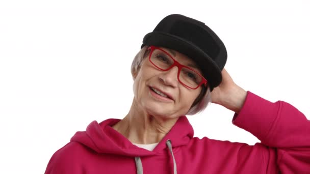 鮮やかさの本質を捉え 65歳以上の女性は 洗練された黒い帽子と純粋な白い背景に設定された赤い眼鏡で補完された 彼女のトレンディな赤いパーカーで幸せを表現しています Rawについて — ストック動画
