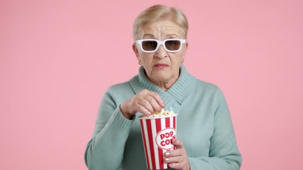 金发迷人的老妇人 带着爆米花坐在纸杯里 带着粉红的背景 望着镜头 屏住呼吸 害怕地看着接下来会发生什么 高质量的4K镜头 — 图库视频影像