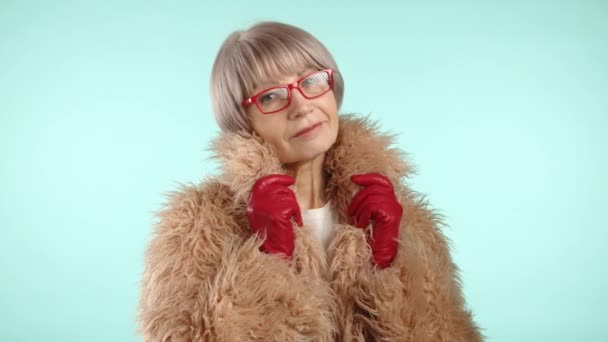 60代の女性は 冬のシックなふわふわのタンニクの毛皮のコートを包み込み 赤い手袋とガラスをパステルブルーの背景と調整しています カメラ Raw — ストック動画