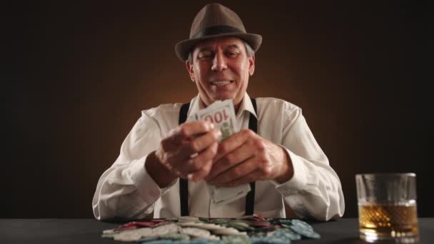 フードラ帽子とポーカーゲームからの現金賞金の積み重ねをカウントし ダークバックドロップに対して近くのウイスキーグラスで楽しむ カメラ Raw — ストック動画
