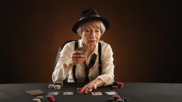 一名65岁的女人穿着黑社会风格的服装 身穿白衬衫 打着宽松的黑色领带 在桌子上发现了扑克牌 她在一间昏暗温暖的房间里仔细审视着自己的手 8K照相机 — 图库视频影像