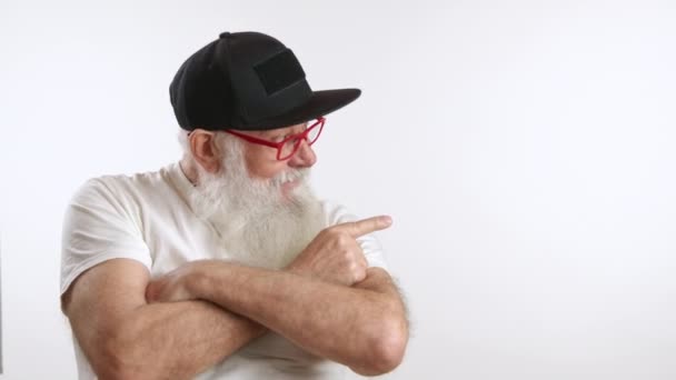 75歳のジョイフルな男性は フレームの空っぽの部分に指を示しています あなたの製品 テスト または他のものを宣伝する 白い背景に白いTシャツ 黒い帽子 赤いメガネ カメラ Raw — ストック動画