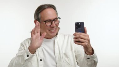 Gözlüklü mutlu bir yaşlı adam bir video görüşmesine katılırken sıcak bir şekilde gülümsüyor, akıllı telefonunu kaldırıyor, neşesi temiz beyaz bir fon karşısında belirginleşiyor. Kamera 8K HAM. 