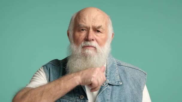 白いひげを持つ高齢者は 柔らかいターコイズの背景に対して 脅威や議論の終わりを意味する彼の手で喉を切るジェスチャーを作っています カメラ Raw — ストック動画