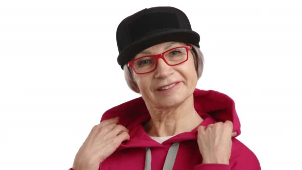 一位超过65岁的令人愉快的女人 身穿红色帽衫 戴着黑色帽子 戴着红色眼镜 与清澈的白色背景形成了鲜明对比 脸上洋溢着欢乐的光芒 8K照相机 — 图库视频影像