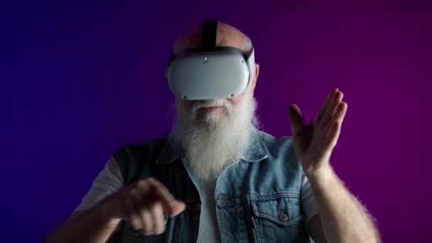 令人惊讶的老人 双手举着虚拟实相耳机 身穿牛仔背心 背景是鲜活的紫色 8K照相机 — 图库视频影像