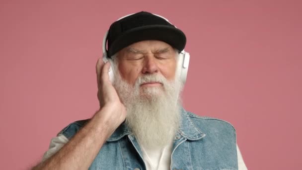 完全な白いひげを持つコンテンツの高齢者は 目を閉じて落ち着いた音楽を楽しんで ピンクの背景にヘッドフォンを着ています カメラ Raw — ストック動画