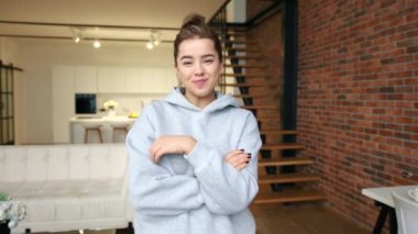 Kendine güvenen genç bir kadın kollarını katlamış gri kapüşonlu bir kapüşonluyla arka planda bir mutfakla modern evinde rahatça poz veriyor. Kamera 8K HAM.