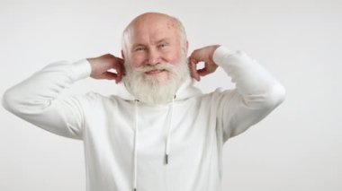 Açık renk sakallı, beyaz kapüşonlu, çekici yaşlı bir adam, kapüşonunu takıyor ve kollarını göğsünde çapraz yapıyor, beyaz bir arka plana gülümsüyor. Kamera 8K HAM. 