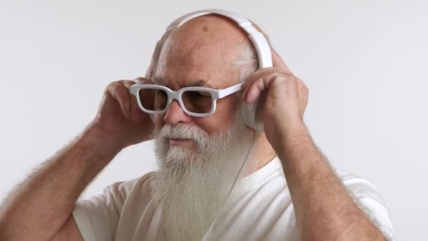 一位留着白胡子的老人正在竖起白色耳机 听他最喜欢的曲子 穿着白衬衫 在简约的白色背景下表现出宁静 8K相机 — 图库视频影像