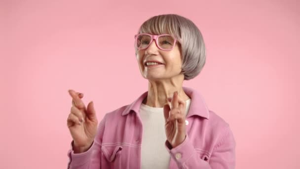 一位优雅的老妇人 带着迷人的笑容 带着时髦的粉色眼镜和配套的夹克 用交叉的手指做了一个热切的手势 在柔和的粉色背景下摆出姿势 8K照相机 — 图库视频影像