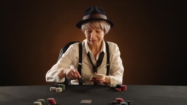 居心地の良い雰囲気の薄暗い部屋で 65歳の女性がギャングの衣装を着て白いシャツと黒い帽子を着て テーブルのチップに囲まれたポーカーカードを扱う笑顔 — ストック動画