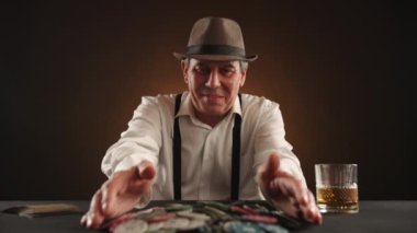 Poker maçını şapkalı ve pantolon askısı olan beyaz gömlekli neşeli bir adam kazandı. Tüm çipleri kendine toplamaktan mutluluk duyar. Yakınlarda yalan kartları ve bir bardak viski var. Kamera 8K HAM.