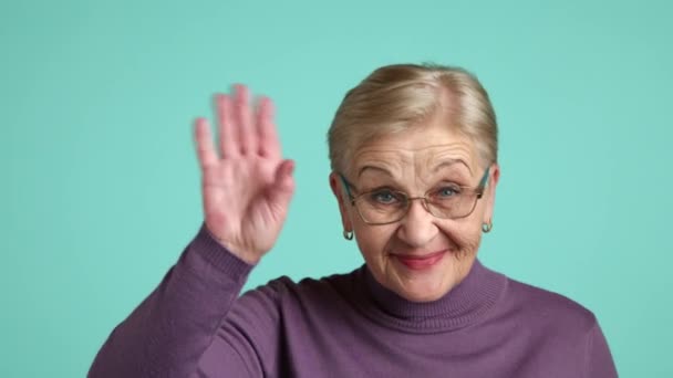 一位快乐的老太太 戴着眼镜 金发碧眼 短短的头发 穿着紫色的高领毛衣 站在蓝色的背景上 挥挥手 微笑着 高质量的4K镜头 — 图库视频影像