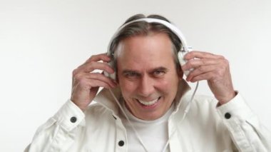 Orta yaşlı bir adamın zevkle beyaz kulaklık taktığı ve mutlu bir şekilde müzik dinlediği parlak bir sahne. Beyaz bir ceket, beyaz bir tişört giyer ve beyaz bir arka planı vardır. Kamera 8K HAM.