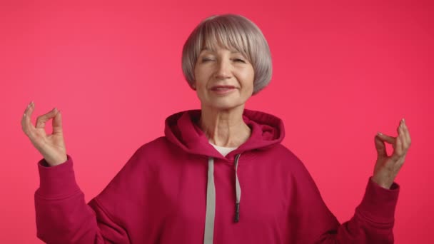 一位面带微笑的老妇人在冥想之后睁开眼睛 这是她的帽衫的栗色 补充了柔和的粉色背景 营造出一种满足和放松的形象 8K照相机 — 图库视频影像