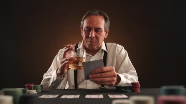 中年の頑丈な男がポーカーをしている 彼はタバコを吸い 片手にカードを握り もう片方には高価なウイスキーのグラスを持っている 男は彼の勝利に自信を持っており 相手に笑顔 Raw — ストック動画