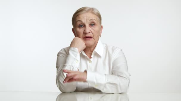 一位60岁至70岁的金发女人 身穿白衬衫 坐在白色的背景上 敲着桌子上的手指 出于误解而伸出手来 高质量的4K镜头 — 图库视频影像