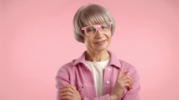 シルバーヘアのスタイリッシュな女性が笑顔でピンクのメガネを調整 平坦な背景に柔らかいピンクのジャケットを着用しています カメラ Raw — ストック動画