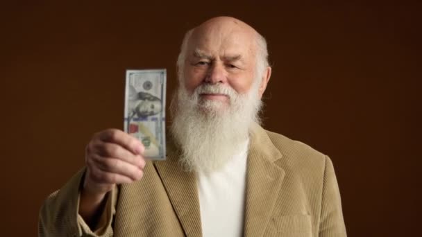 一位满头白胡子的快乐的老年绅士骄傲地举起大拇指 穿着经典的米黄色夹克 背景浓密的棕色衬托下 在表示认可的同时 自豪地出示钞票 — 图库视频影像