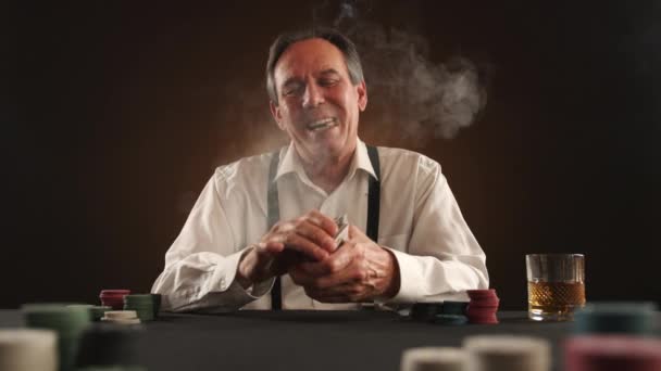 ポーカーテーブルに座ってタバコを吸う高齢者 テーブルの上 高価なウイスキー チップ カード 男はサスペンサー付きの白いシャツのギャングのように見えます — ストック動画