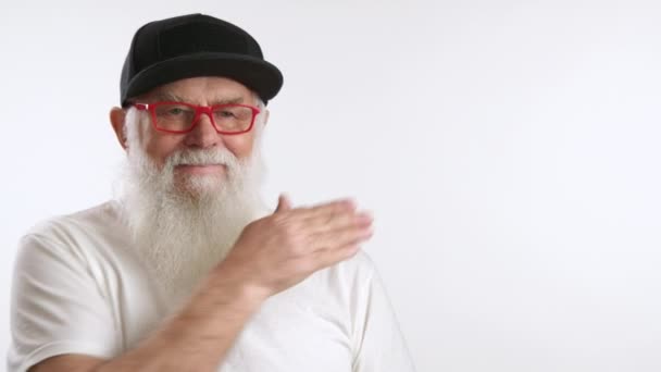 テキスト配置 ロゴの画面の右側を指す大きな灰色のひげを持つスタイリッシュな祖父 白いTシャツ 黒い帽子 赤いメガネを着ている男 ホワイトバック — ストック動画