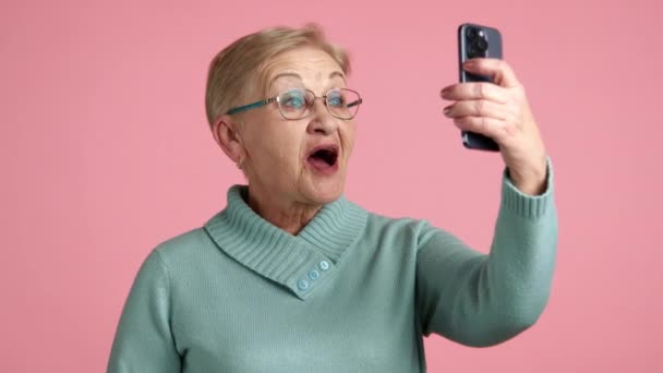 素敵なブロンドの高齢女性は 携帯電話でテレコンフェンシングを見て 非常に幸せなインターロケーター 手を振ってピンクの背景の上に立って 高品質の4K映像 — ストック動画