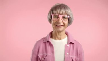 Işıldayan bir gülümsemesi olan, pembe ceketli ve gözlüklü neşeli yaşlı bir kadın yumuşak pembe bir arka plana karşı başını sallıyor. Kamera 8K HAM. 