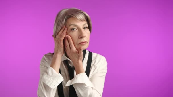 一位老年妇女正在细腻地摸着她的脸颊 看着相机 用生动的紫色背景展现了一种细心的护肤习惯 — 图库视频影像