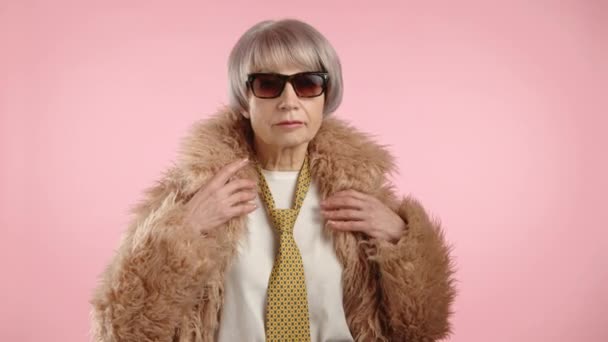 时髦的老妇人 戴着太阳镜 穿着时髦的假皮毛外套 自信地摆姿势 在柔和的粉色背景下体现出优雅和时尚 8K照相机 — 图库视频影像