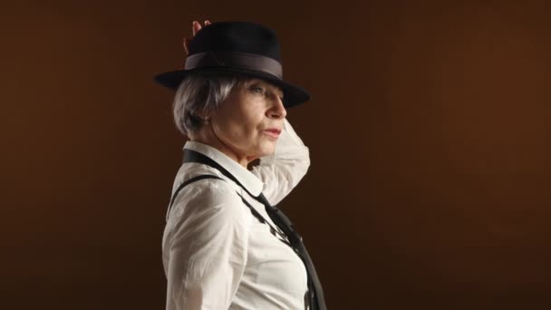 一位六十多岁的优雅的老年妇女 穿着时髦的法兰西斯帽 打着领带 在深棕色背景下流露着风格和经验 理想的成熟时尚主题 — 图库视频影像