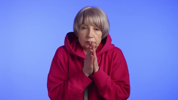 美丽的老妇人因寒冷而发抖 她用红色的帽衫和蓝色的单色背景擦拭着冰冷的手 8K照相机 — 图库视频影像