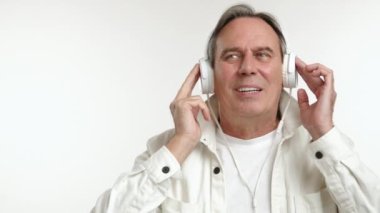 Beyaz ceketli neşeli, olgun bir adam, kulaklıkla müzik eşliğinde kendini kaybediyor, yüzü mutluluk dolu bir gülümsemeyle aydınlanıyor, beyaz bir altyapısı var. Kamera 8K HAM. 