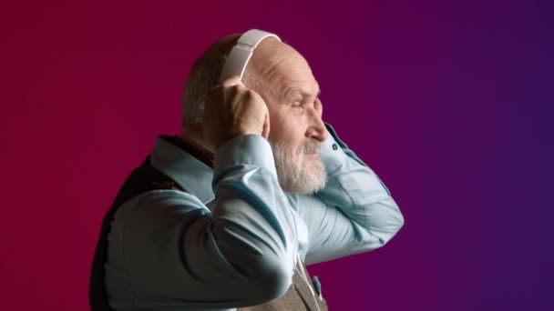 明るいシャツとベストを着た高齢者がヘッドフォンを着て 静かな音楽を楽しんで目を閉じ 活気に満ちた紫色の背景に囲まれています 高品質の4K映像 — ストック動画