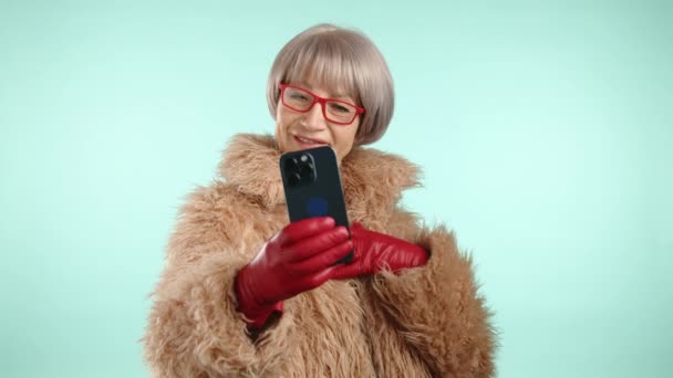 时尚的老年女性 穿着橙色的生态皮毛外套 戴着红色手套 戴着红色眼镜 留着短发 在智能手机上自拍 蓝色背景 8K照相机 — 图库视频影像