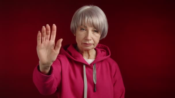 一位年事已高的妇女举起她的手 用她的手掌做了一个停止的手势 摇摇头表示不赞成 穿着红色的帽衫 帽檐与红色的背景相对应 8K照相机 — 图库视频影像