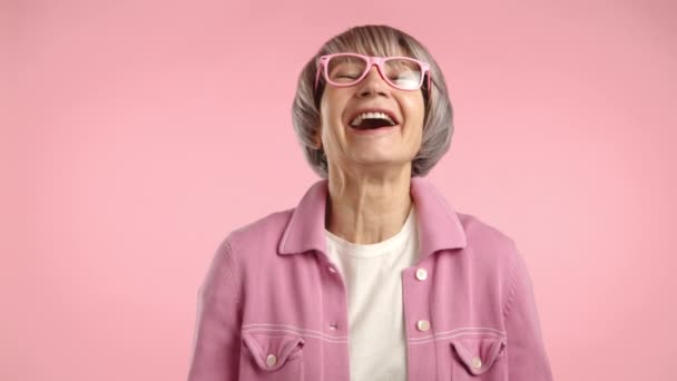人生に満ちた高齢女性 スタイリッシュなピンクグラスで心のこもった笑いを楽しみ 調整ピンクの背景に対してポジティブさと喜びを放つ — ストック動画
